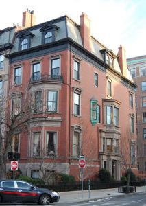 A large historic brick building with a sloped roof, featuring multiple windows and balconies, located at the corner of a street. The façade includes a green decorative element and nearby traffic signs.